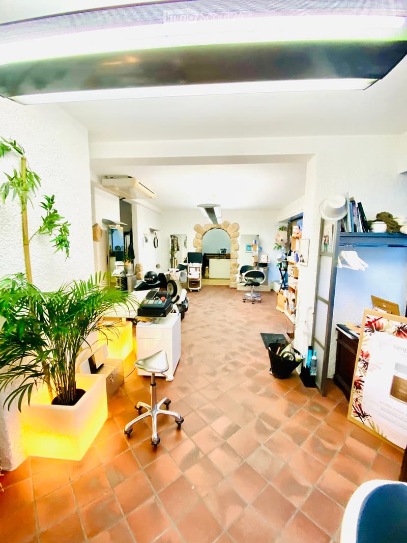 Bright, open room with terracotta tiled floor, white walls, and modern furniture including a chair, stool, and plant. A mirror and shelves are visible in the background.