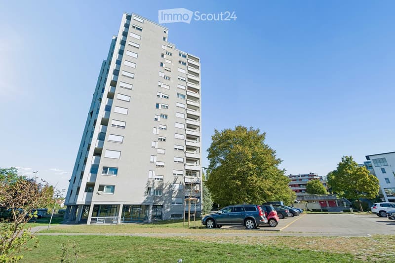 Modern multi-story apartment building with balconies, surrounded by trees and a parking lot with several cars.