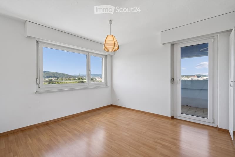 Empty room with wooden flooring, white walls, large windows, and a sliding glass door leading to a balcony.