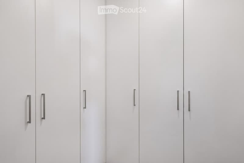 A completely empty room with white walls and white built-in wardrobes with silver handles.