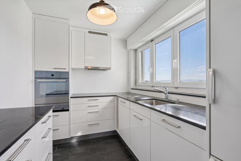 Modern kitchen with white cabinets, black countertops, stainless steel sink, and built-in oven. Features large windows and a pendant light fixture.