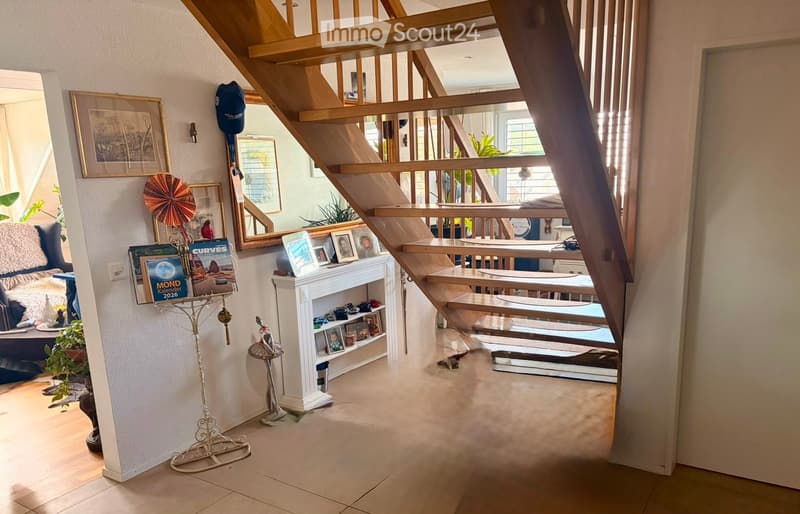 A well-lit hallway with a wooden staircase leading to an upper level. The area features a cozy blue sofa with a fluffy throw, a small table with a calendar, a shelf with picture frames and decorative items, and a mirror on the wall. The floor is tiled, an