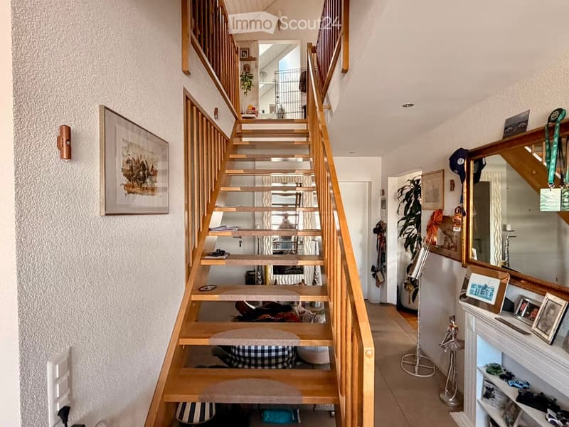 Wooden staircase with beige carpeted steps, wooden railings, and a small landing area. The hallway features a framed picture on the left wall, a mirror on the right, and a small shelf with framed photos and decorative items.