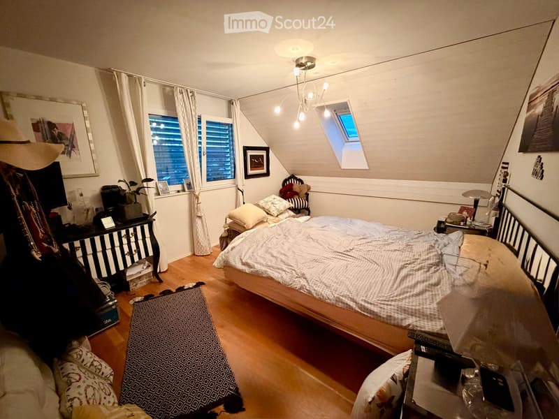 Cozy attic bedroom with wooden floors, slanted ceilings, and large windows. Features a double bed with patterned bedding, a nightstand with a lamp, a dresser with a mirror, and a small seating area with a chair and plant.