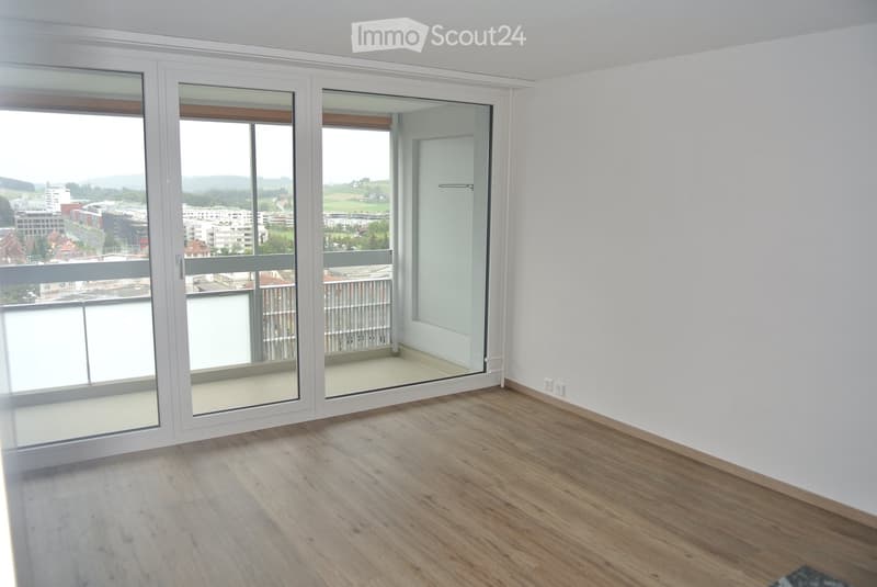 Empty room with wooden flooring, white walls, large sliding glass doors leading to a balcony, and a view of the city and landscape.