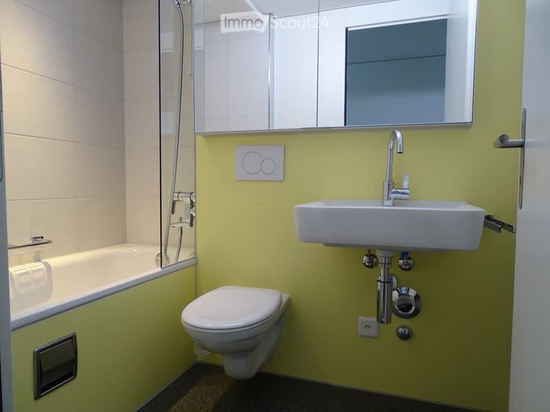 Modern bathroom with a bathtub, toilet, sink, and mirror. The walls are tiled in light beige and bright green, and the floor is dark.