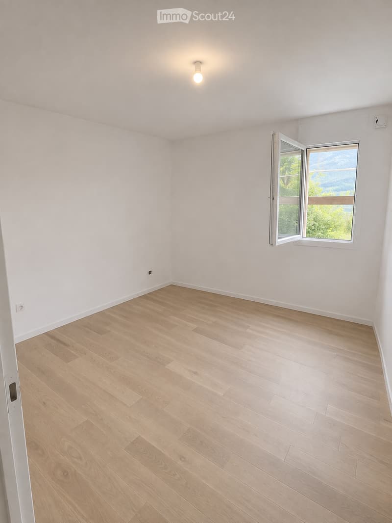 Empty room with light wooden flooring and white walls, featuring a large window with a scenic view and a ceiling light fixture.