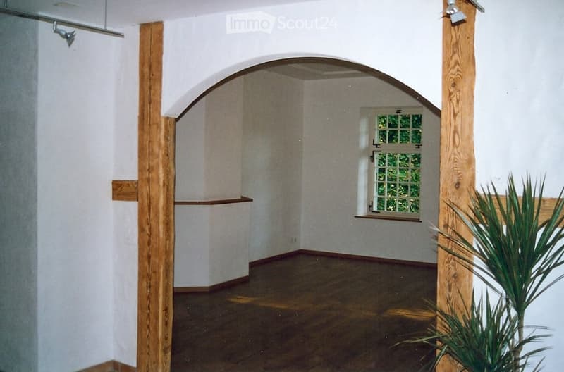 Empty room with wooden floor, white walls, wooden pillars, arched doorway, small window, and a plant.