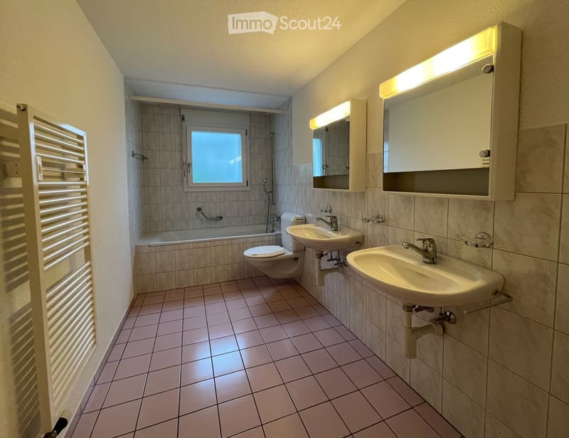 Modern bathroom with two sinks, toilet, bathtub, and built-in mirrored cabinets with lighting. Features tiled walls and floor, a window above the bathtub, and a radiator on the left side.
