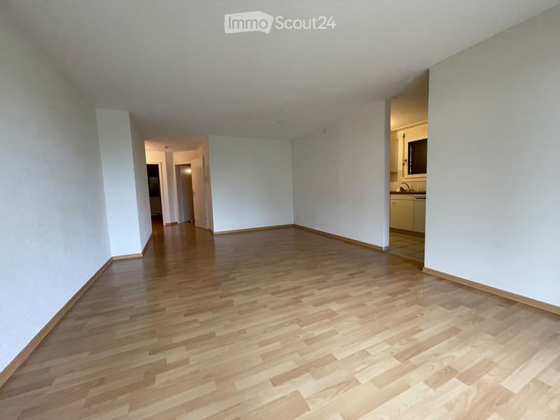 Empty room with wooden flooring and white walls, featuring an open doorway leading to another room and a small kitchen area visible in the background.