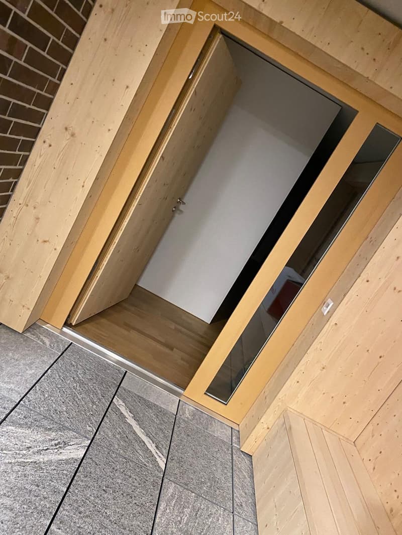 A modern hallway with wooden flooring and a glass door leading to another room. The walls are painted white, and the ceiling is made of light wood.