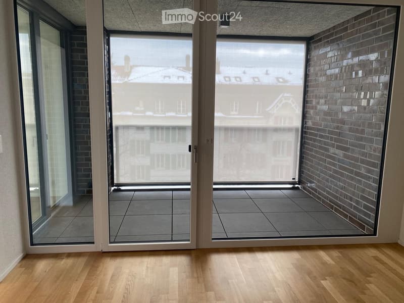 Empty room with wooden flooring, large glass doors leading to a balcony with tiled floor and brick wall.