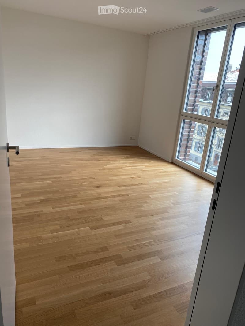 Empty room with light wooden flooring and white walls, featuring large windows with a view of the street.