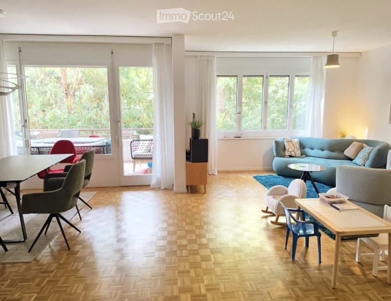 Bright living room with wooden flooring, large windows, and a balcony. Features a blue sofa, coffee table, dining area with chairs, and a small plant on a side table.