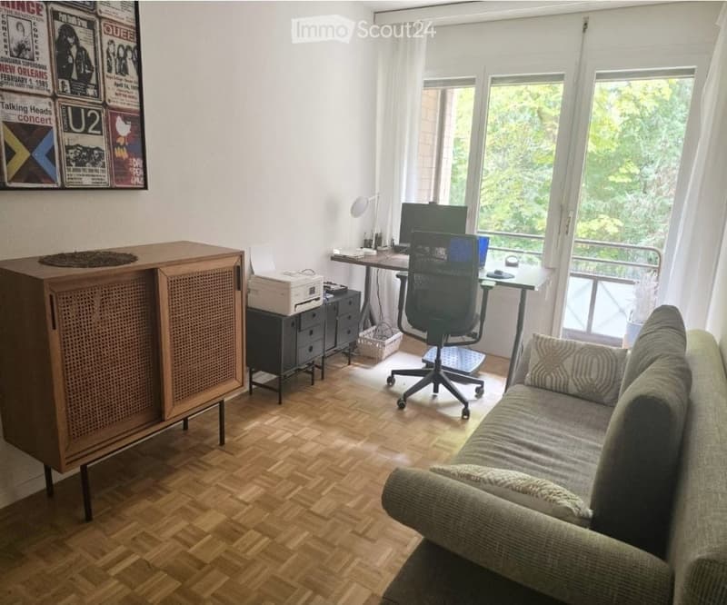 A modern, multi-functional room with a wooden herringbone floor, a grey sofa, a black office chair, a desk with a computer, a wooden cabinet, and a printer. Large windows and a balcony offer natural light and an outdoor view.