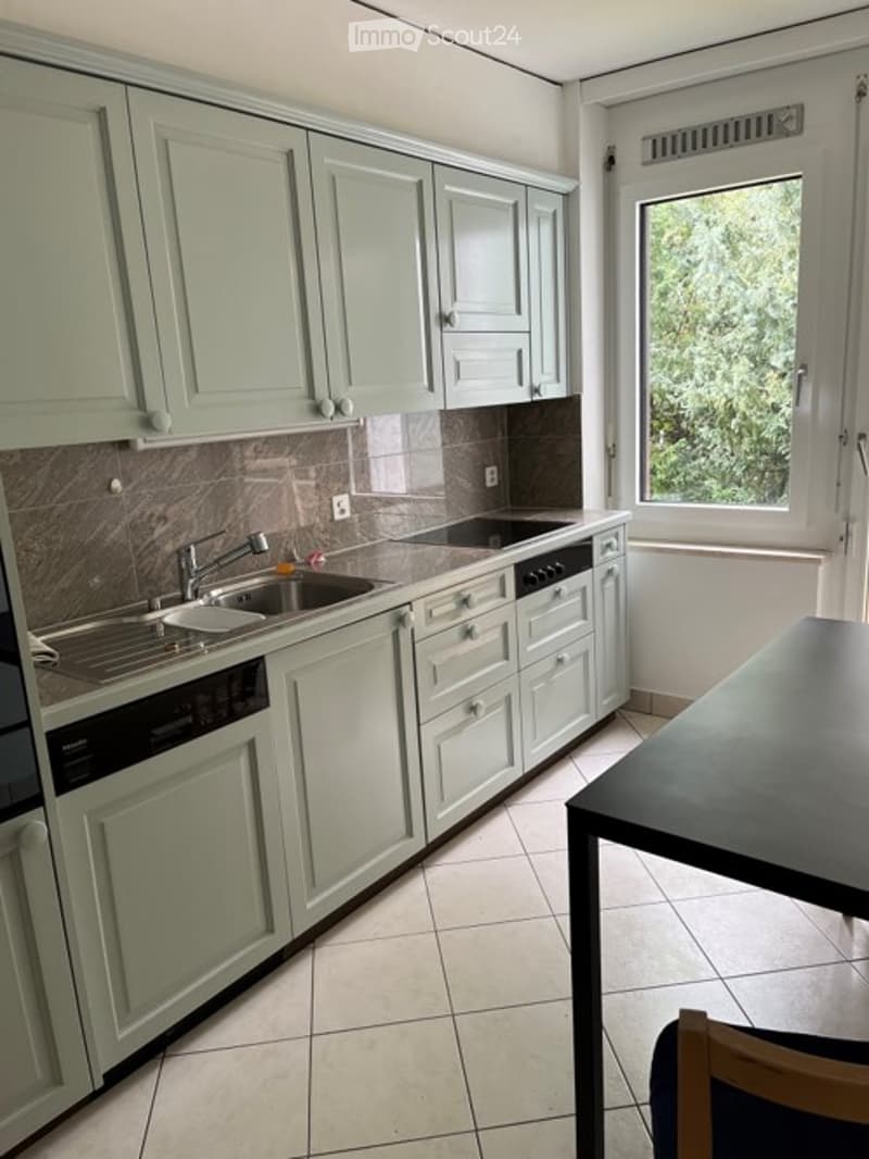 Modern kitchen with light green cabinets, stainless steel sink, dishwasher, and a small dining area with a table and chair.