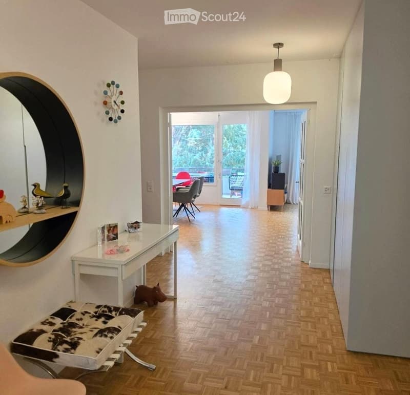 Modern hallway with wooden flooring, white walls, a hanging light fixture, and a round mirror on the wall. A small table with decorative items and a bench are visible, leading to an open area with chairs and a table.