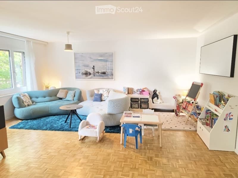 Bright living room with wooden flooring, large windows, a blue sectional sofa, a small coffee table, a child's chair, a small table with chairs, a bookshelf, and a large screen on the wall.