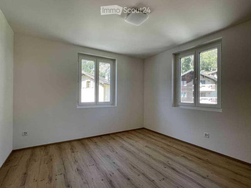 Empty room with wooden flooring, white walls, and two windows offering views of neighboring buildings.