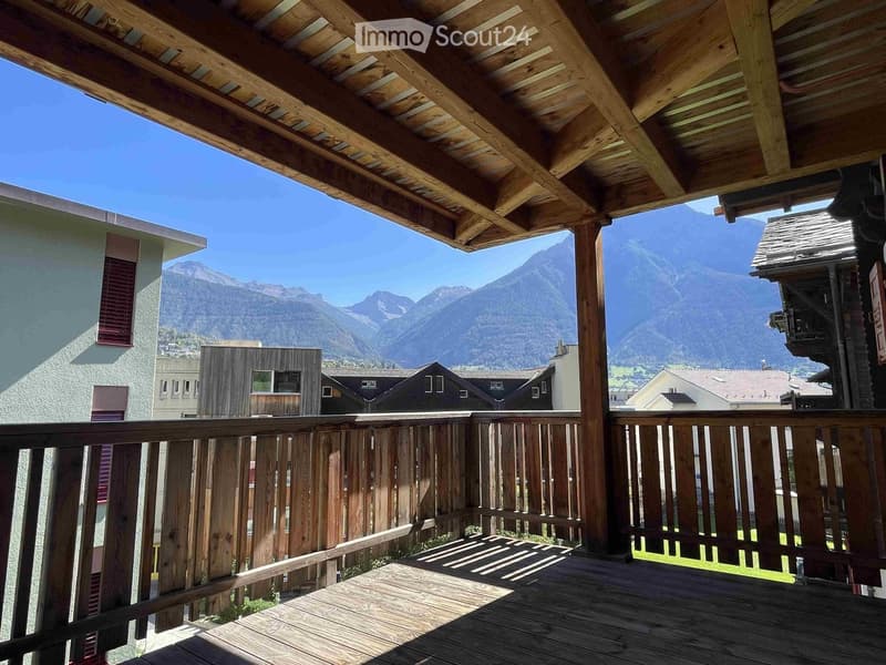 Wooden balcony with a railing, overlooking a scenic mountain view and neighboring houses.
