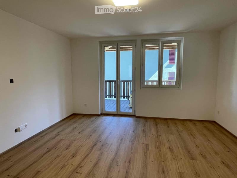 Empty room with wooden flooring, white walls, and large windows with a sliding door leading to a balcony.