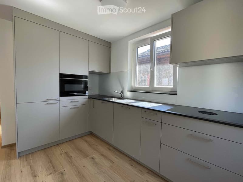 Modern kitchen with grey cabinets, a black countertop, a sink, and an oven. The floor is wooden, and there is a window letting in natural light.