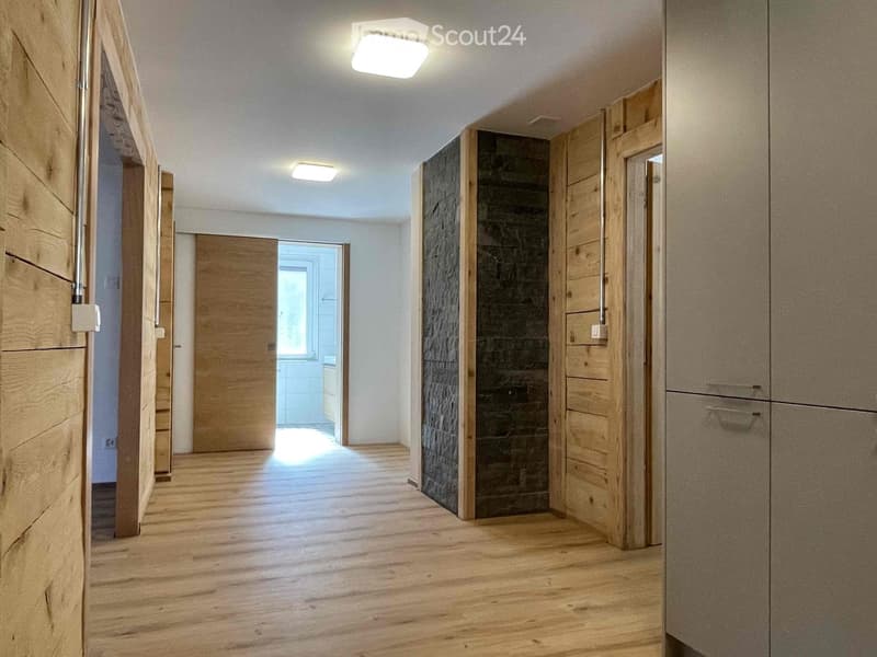 Empty hallway with wooden flooring, white walls, and recessed lighting. Features include a partially open door leading to another room and built-in wooden cabinetry on one side.