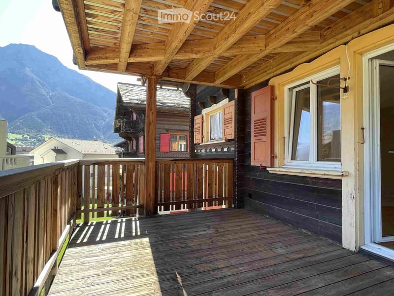 Wooden balcony with railing, overlooking a mountainous landscape with neighboring houses and wooden shutters.