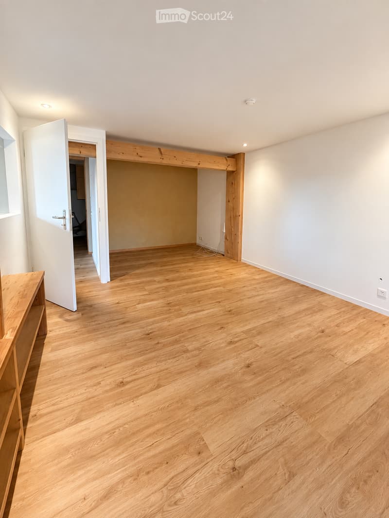 Empty room with light wooden flooring, white walls, and a partially open door leading to another room. The space is clean and unfurnished.