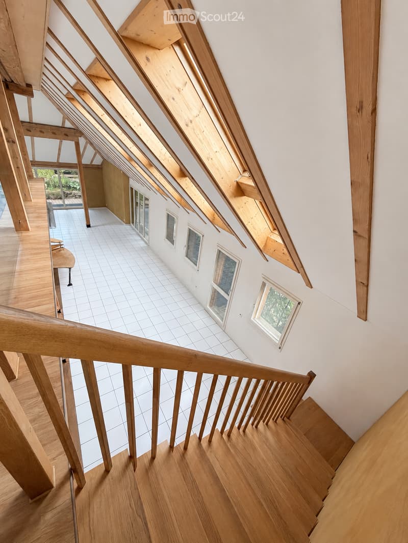 Spacious hallway with wooden staircase, white tiled floor, and large windows allowing natural light. The ceiling features exposed wooden beams and skylights.