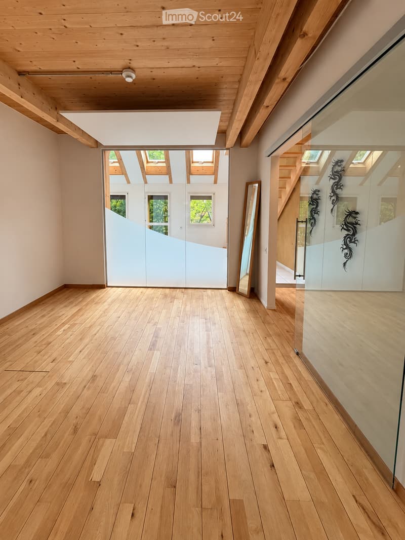 Empty room with light wooden flooring, white walls, and a large glass door leading to another room. The ceiling is exposed wooden beams, and there is a mirror on the wall.