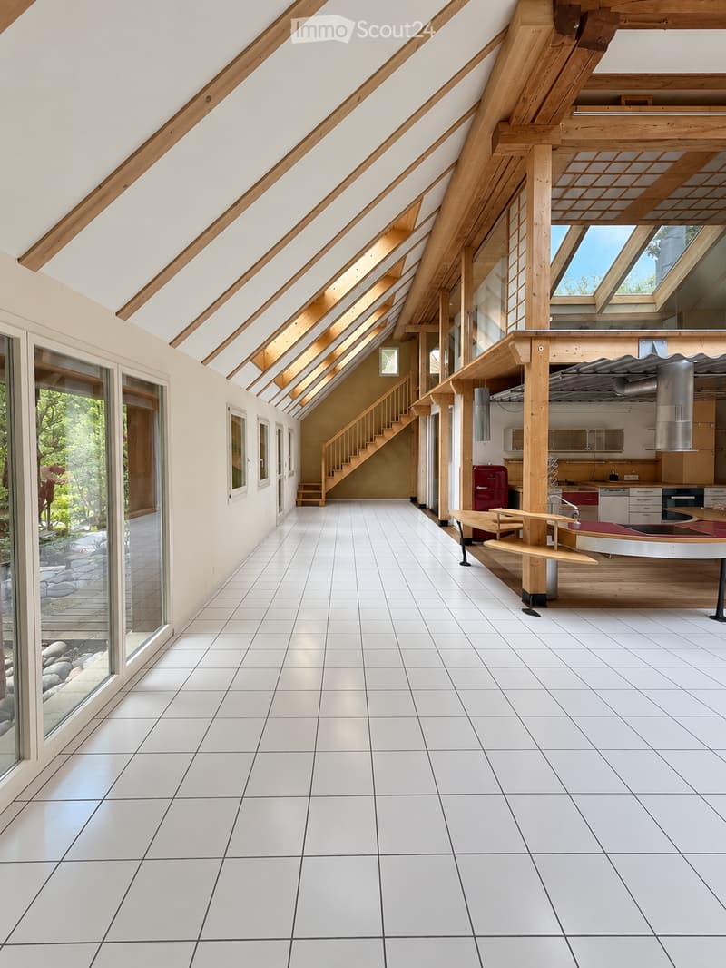 Bright, airy corridor with white tiled flooring, wooden beams, and large windows. Features a small seating area with a table and stools, a staircase leading upwards, and a kitchen area with a refrigerator and island.