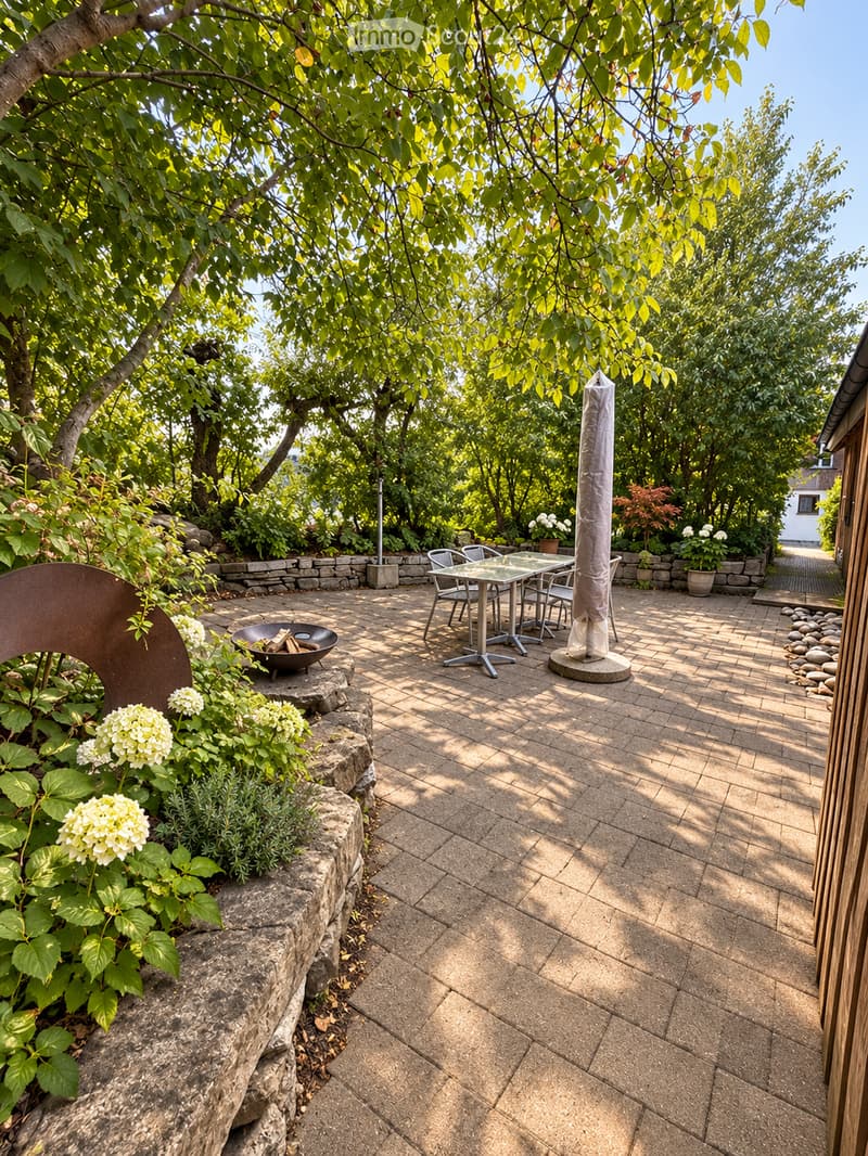 A well-maintained garden patio with a stone-paved floor, a glass-top table with four chairs, a fire pit, and a tall umbrella. The area is surrounded by lush greenery and flowering plants.
