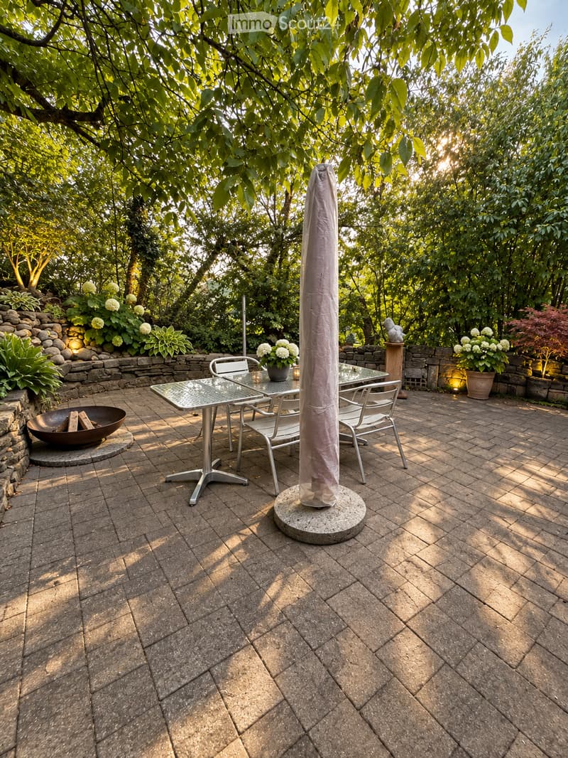 A well-maintained outdoor patio with a brick floor, a glass-top table, four metal chairs, a fire pit, and a covered umbrella pole. The area is surrounded by lush greenery and a stone wall with potted plants.