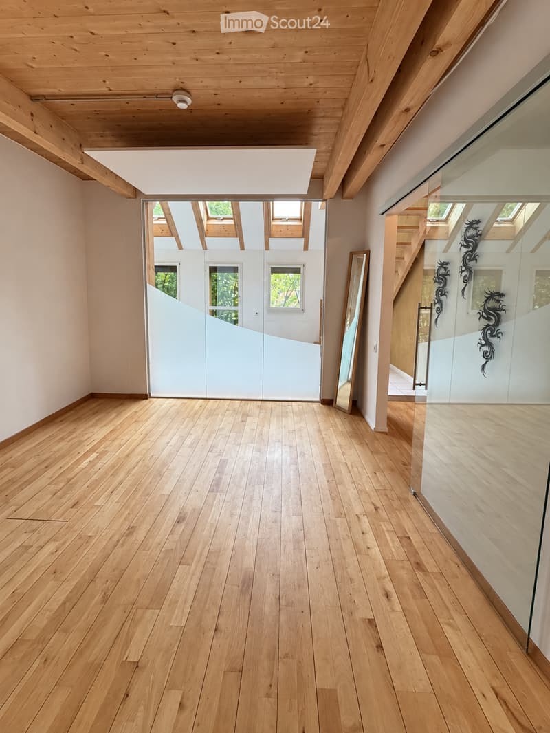 Empty hallway with light wooden flooring, white walls, and large glass partitions leading to another room. The ceiling features exposed wooden beams and skylights.