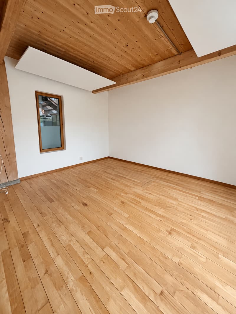 Empty room with light wooden flooring, white walls, and a slanted wooden ceiling. A single window with a wooden frame is visible on one wall.
