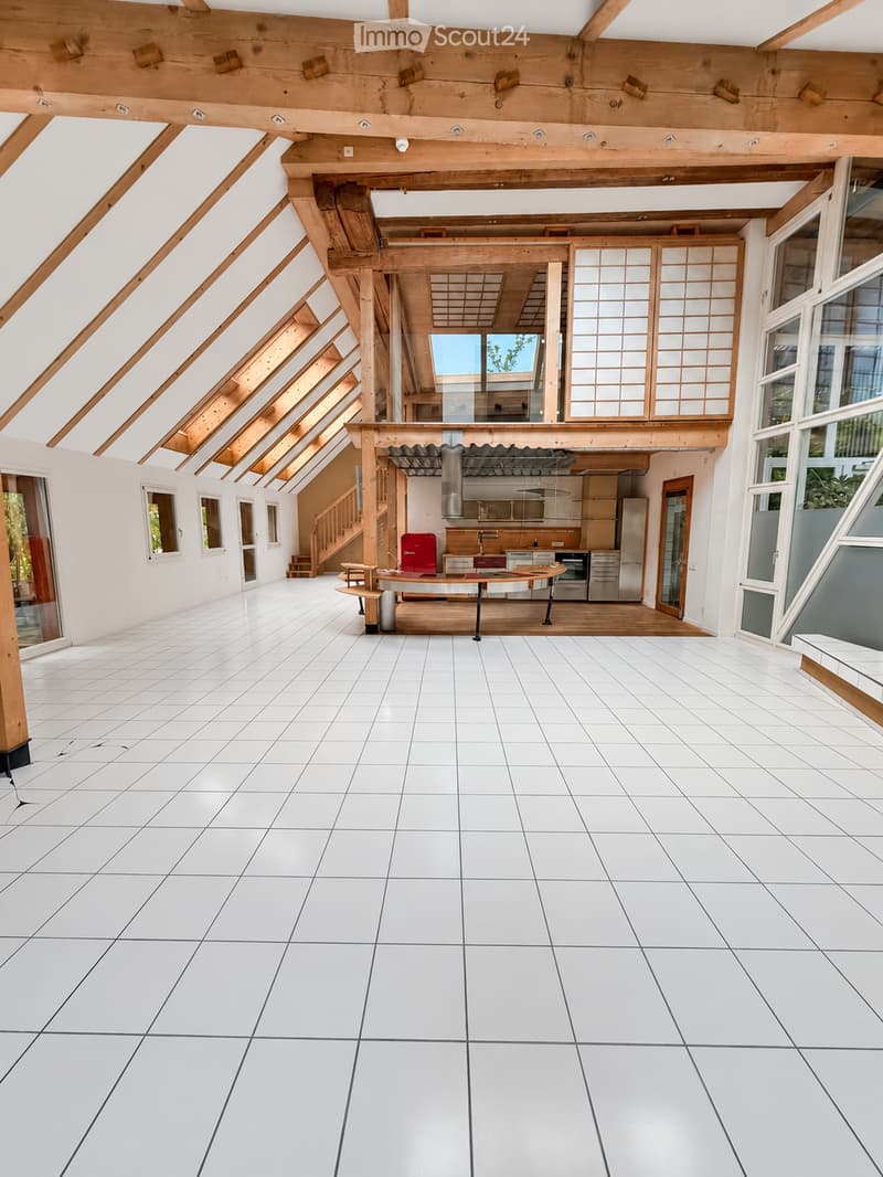 Open, airy space with white tiled floor and exposed wooden beams. Kitchen area visible in the background with cabinets and appliances.