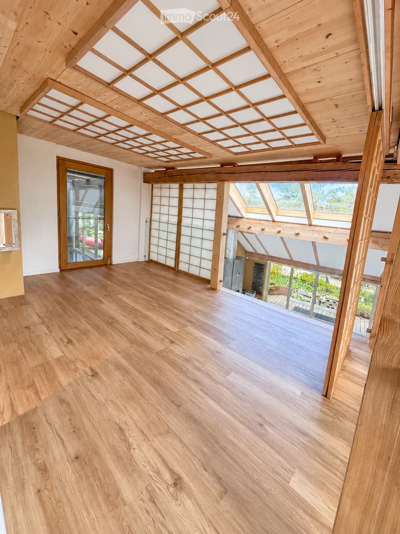 Open, airy room with wooden flooring and exposed wooden beams on the ceiling. Large glass doors lead to an outdoor area, and there are large windows providing natural light.