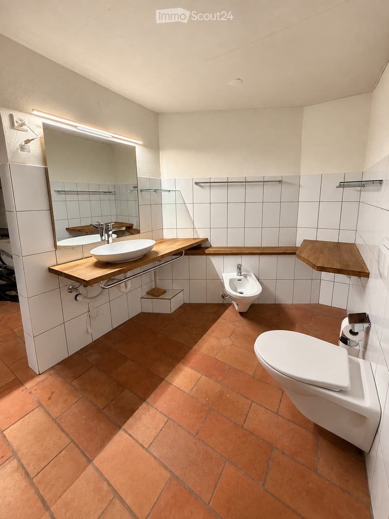 Modern bathroom with white tiles, wooden shelves, a sink, toilet, bidet, and a large mirror with built-in lighting.