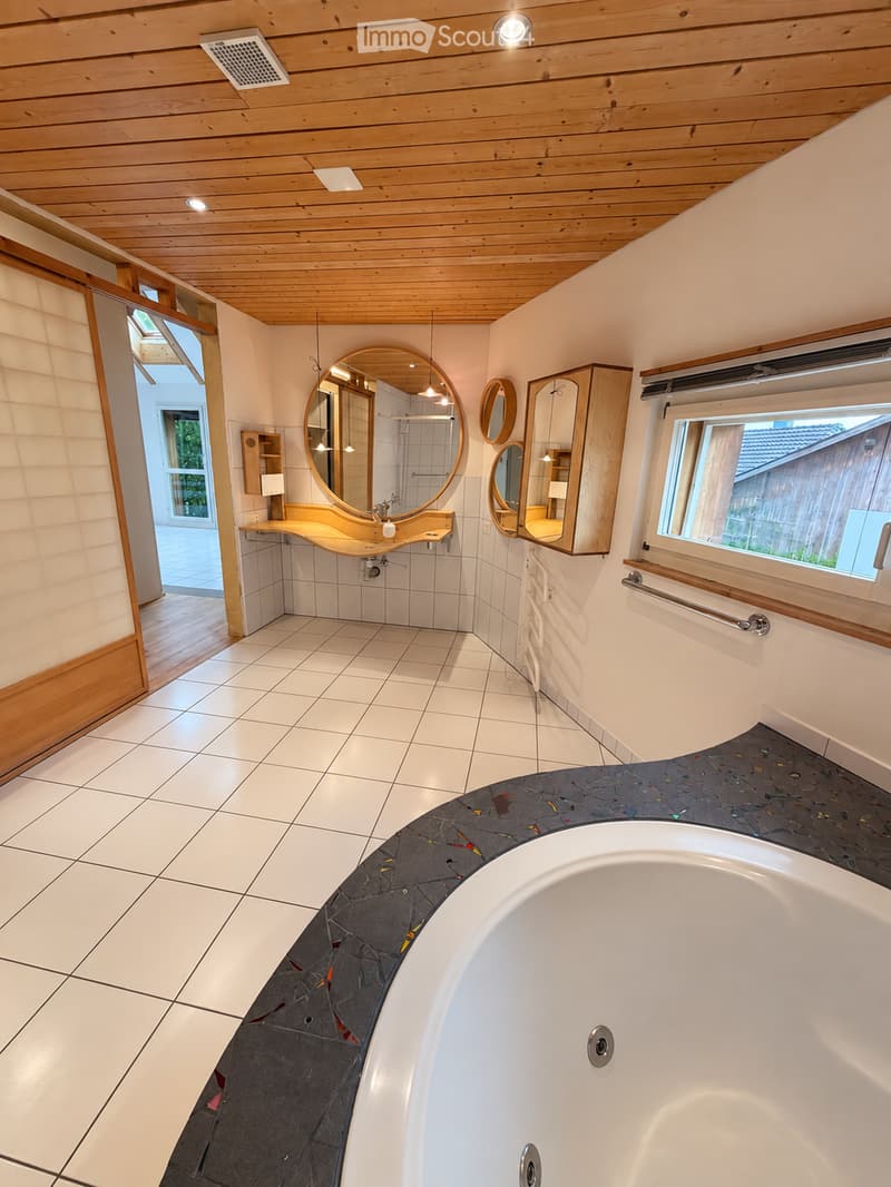 Modern bathroom with white tiled walls and floor, a large bathtub, a sink with a round mirror, and wooden cabinetry.