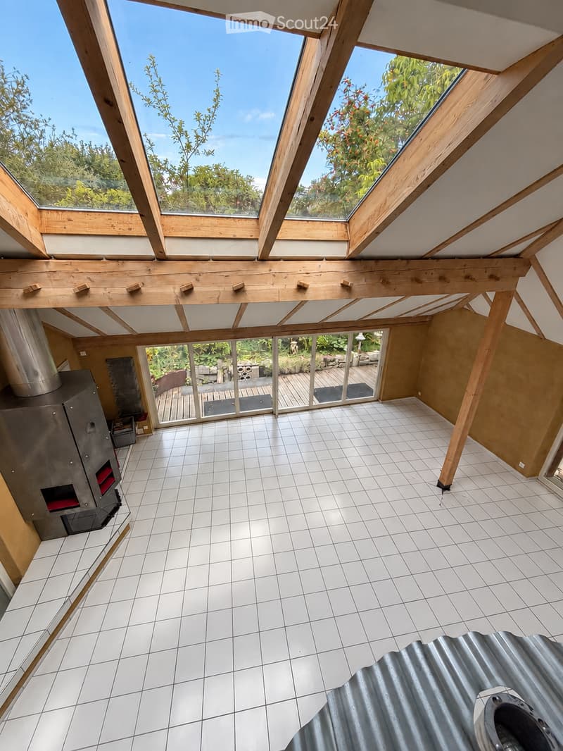Bright, open room with white tiled floor, wooden beams, skylights, and large glass doors leading to an outdoor deck. The space is empty with no furniture.