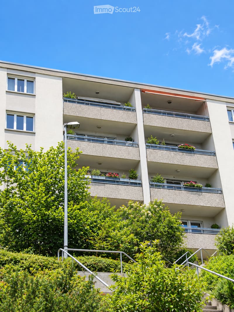 Multi-story residential building with balconies, windows, and greenery in front.