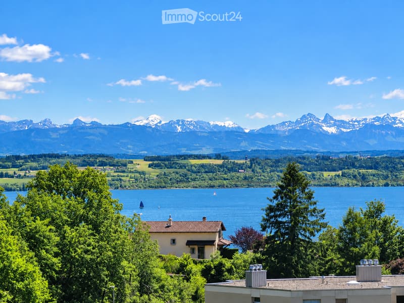 A picturesque view of a house nestled among lush greenery, overlooking a serene lake with sailboats, surrounded by rolling hills and snow-capped mountains under a clear blue sky.