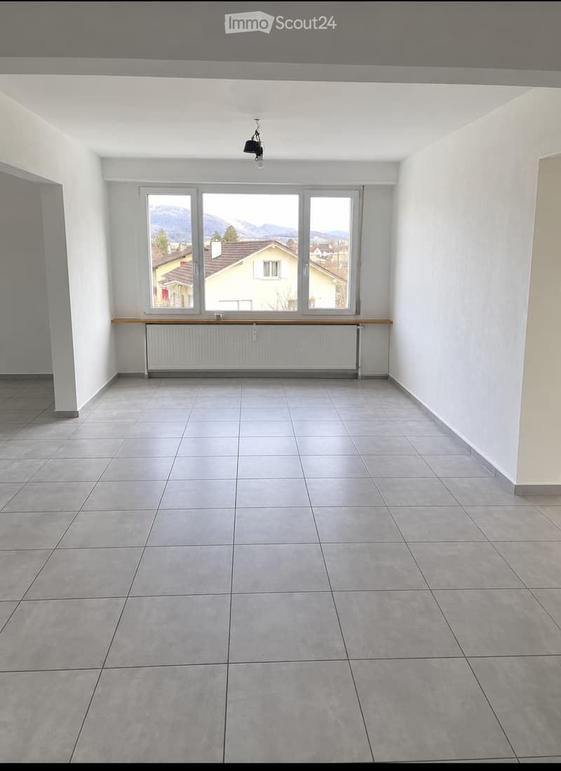 Empty room with tiled floor, white walls, large windows offering a view of mountains and houses, radiator, and a small wooden shelf.