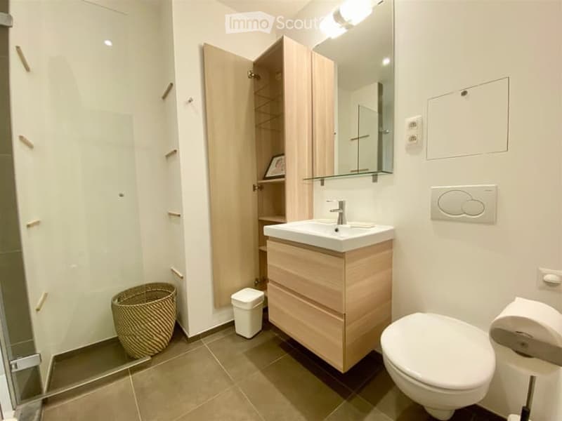 Modern bathroom with a white sink, toilet, mirror, wooden cabinet, wicker basket, and a small trash can. The walls are white, and the floor is tiled in a warm beige color.