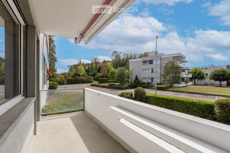 Balcony with white railing, glass screen, and modern design, overlooking a landscaped area with trees, shrubs, and a modern building in the background.