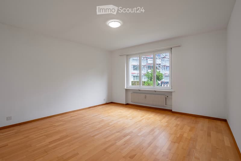 Empty room with wooden flooring, white walls, large window with blinds, radiator, and a ceiling light fixture.