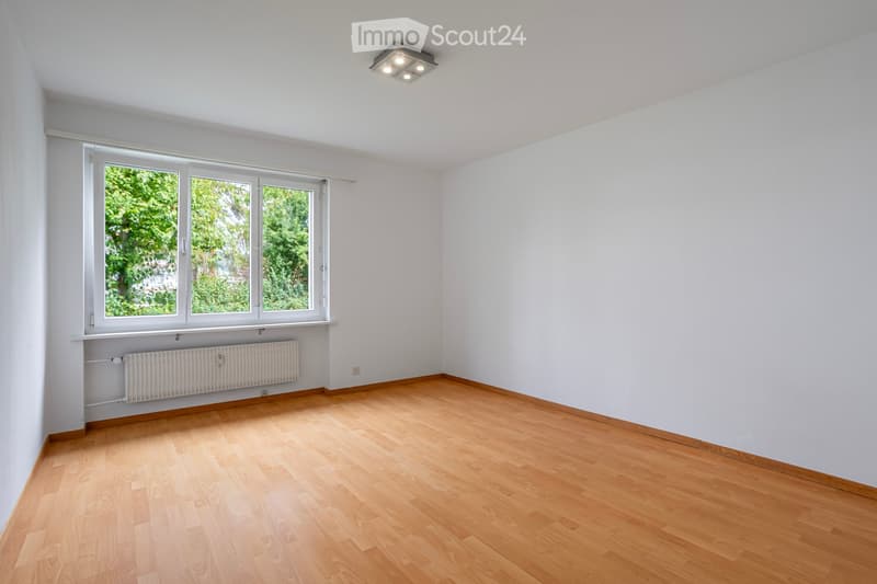 Empty room with light wooden flooring, white walls, large windows with a view of greenery, and a radiator below the windows.
