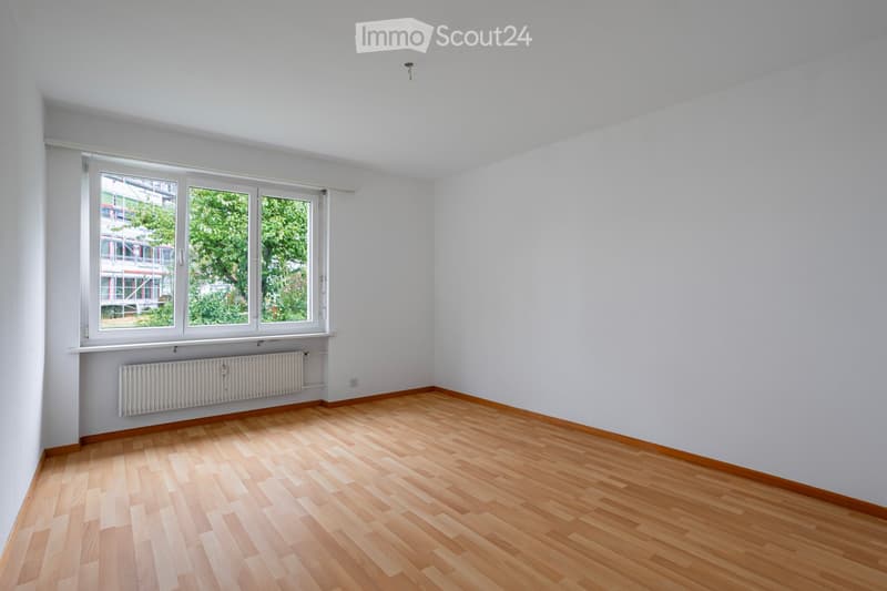 Empty room with light wooden flooring, white walls, and large windows showing greenery outside. Radiator under the windows.