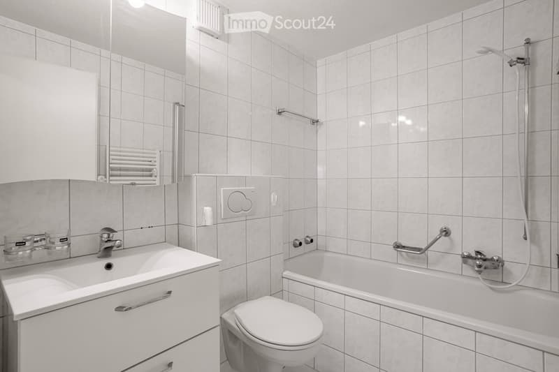 Modern bathroom with white tiled walls and floor, featuring a sink with a mirror, a bathtub with a handheld shower, and a toilet. The sink has a modern faucet and two cups on the counter.